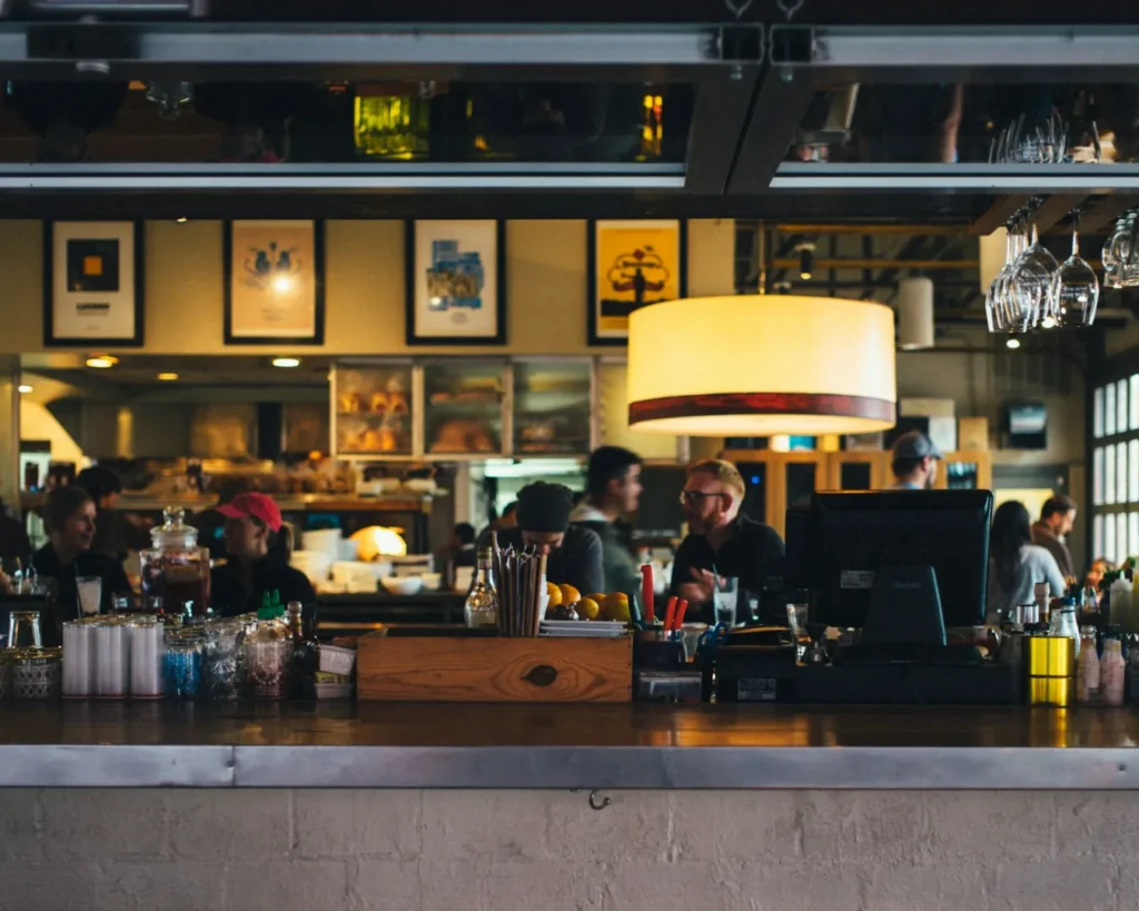 A cozy café with a vibrant atmosphere. People sit and chat at a bar adorned with jars and bottles, while framed art decorates the wall. Warm lighting.
