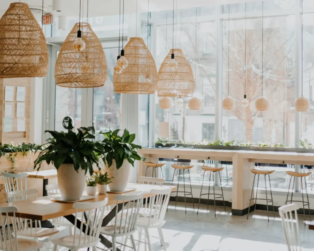 Bright cafe interior with large windows, wooden furniture, and wicker pendant lights. Potted plants on tables create a fresh, inviting atmosphere.