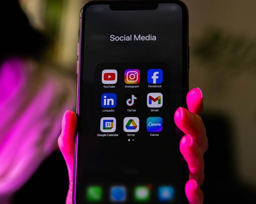 A close-up of a smartphone screen labeled Social Media, showing app icons for YouTube, Instagram, Facebook, LinkedIn, TikTok, and Gmail, held by a hand with bright pink nails