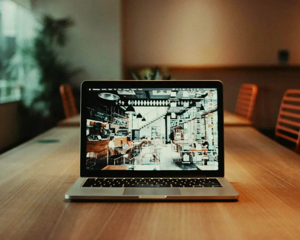 Laptop on a wooden table displaying a lively cafe with people and modern decor. The surrounding room is softly lit, creating a warm atmosphere.