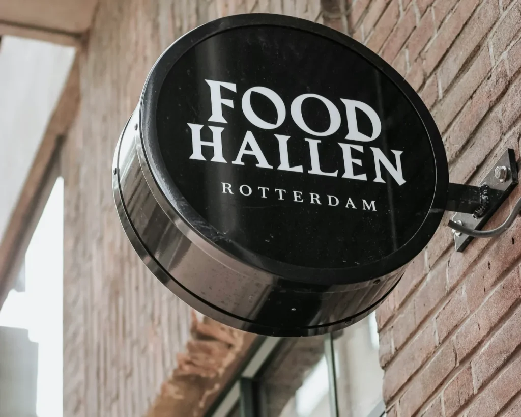 Round black sign reading "Food Hallen Rotterdam" mounted on a brick wall. The sign contrasts with the light bricks, evoking a modern and inviting atmosphere.
