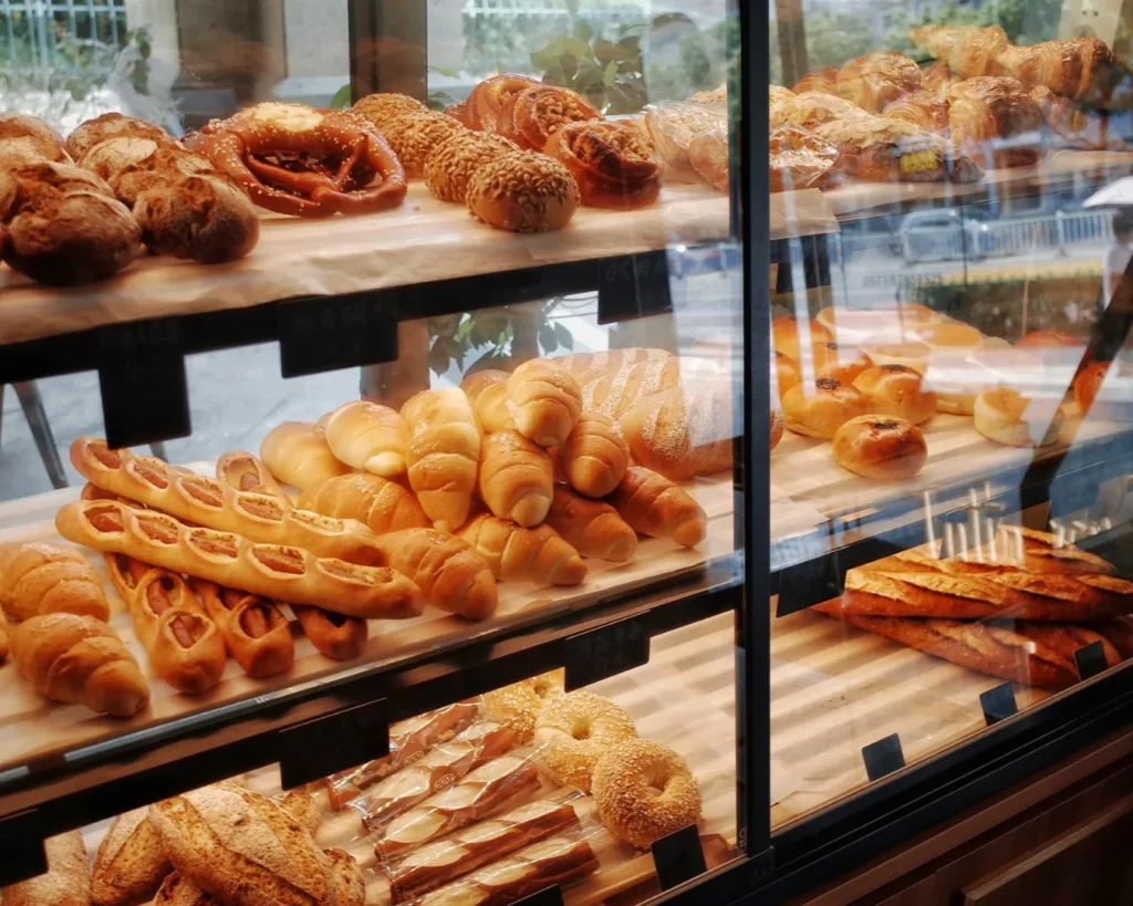 Bakery display with various breads and pastries, including pretzels, baguettes, and croissants, arranged on wooden shelves in warm, inviting light.