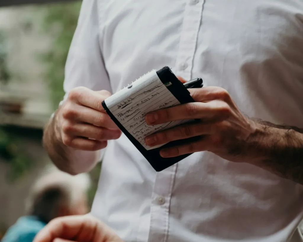 A person in a white shirt holds a notepad and pen, taking an order. The setting appears to be a restaurant with a blurred, leafy background.