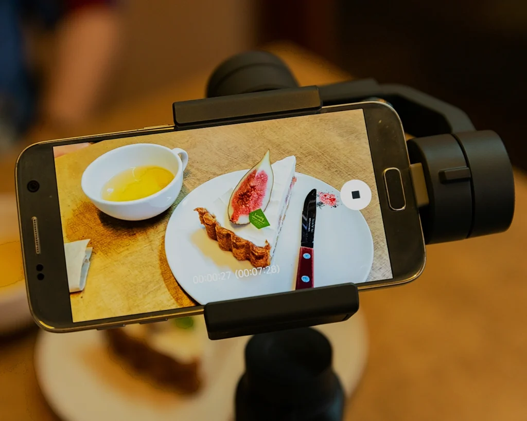 Smartphone on a tripod records a plate with a fig slice, pastry, and knife, beside a cup of yellow liquid, capturing a casual, cozy dining scene.