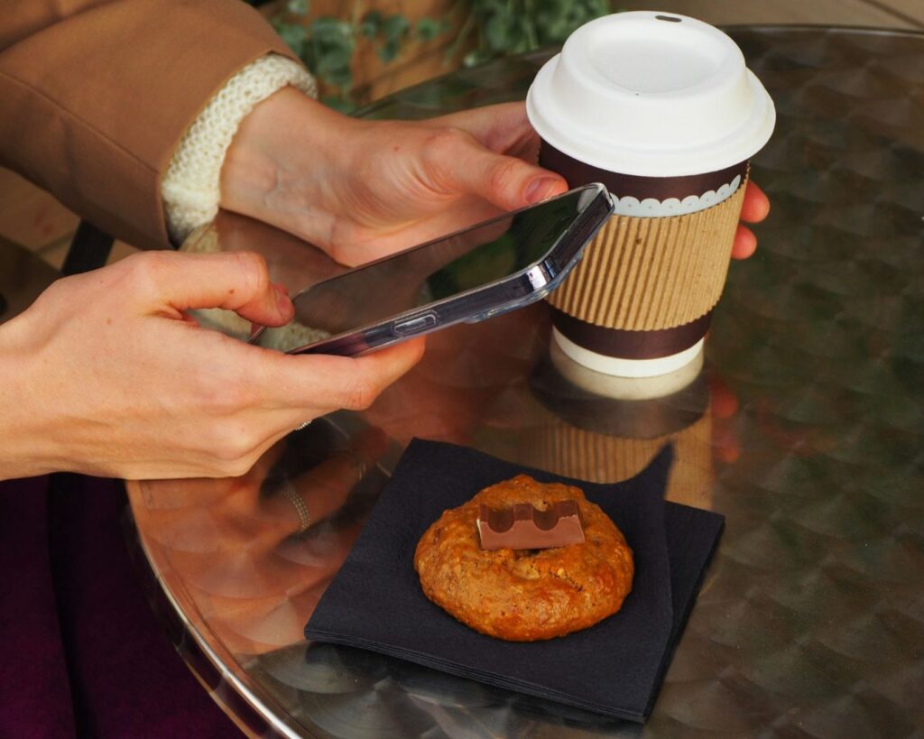 A person holds a smartphone and a coffee cup on a metal table, with a chocolate-topped cookie on a napkin, suggesting a cozy cafe scene.