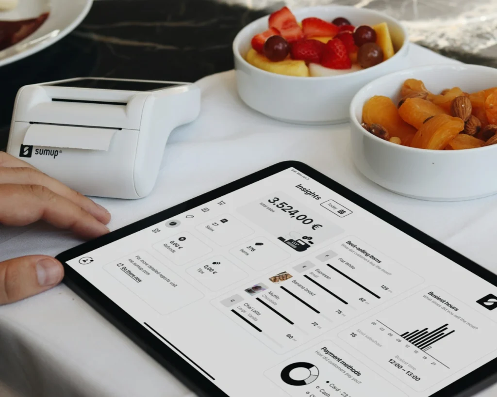 A tablet displays financial data with a payment terminal beside it. Nearby are bowls of fresh fruit and nuts, suggesting a casual dining setting.