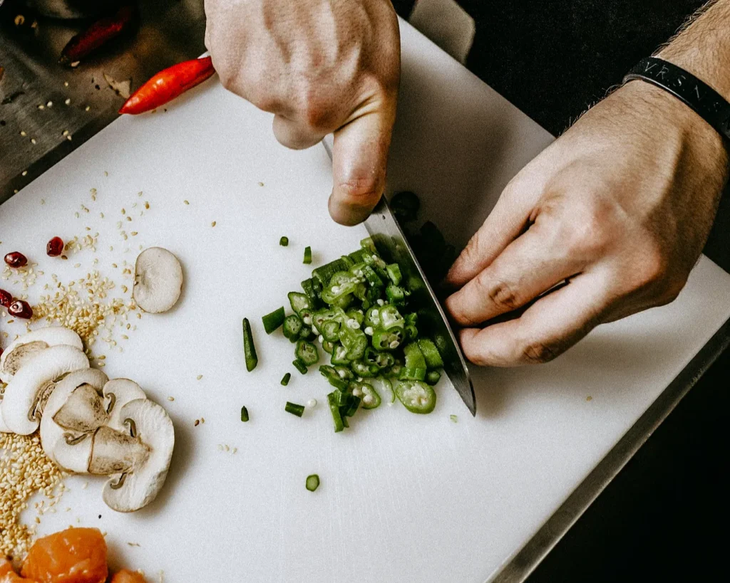 Hands chopping green okra on a white cutting board surrounded by sliced mushrooms, sesame seeds, and red chili peppers, conveying a cooking scene.