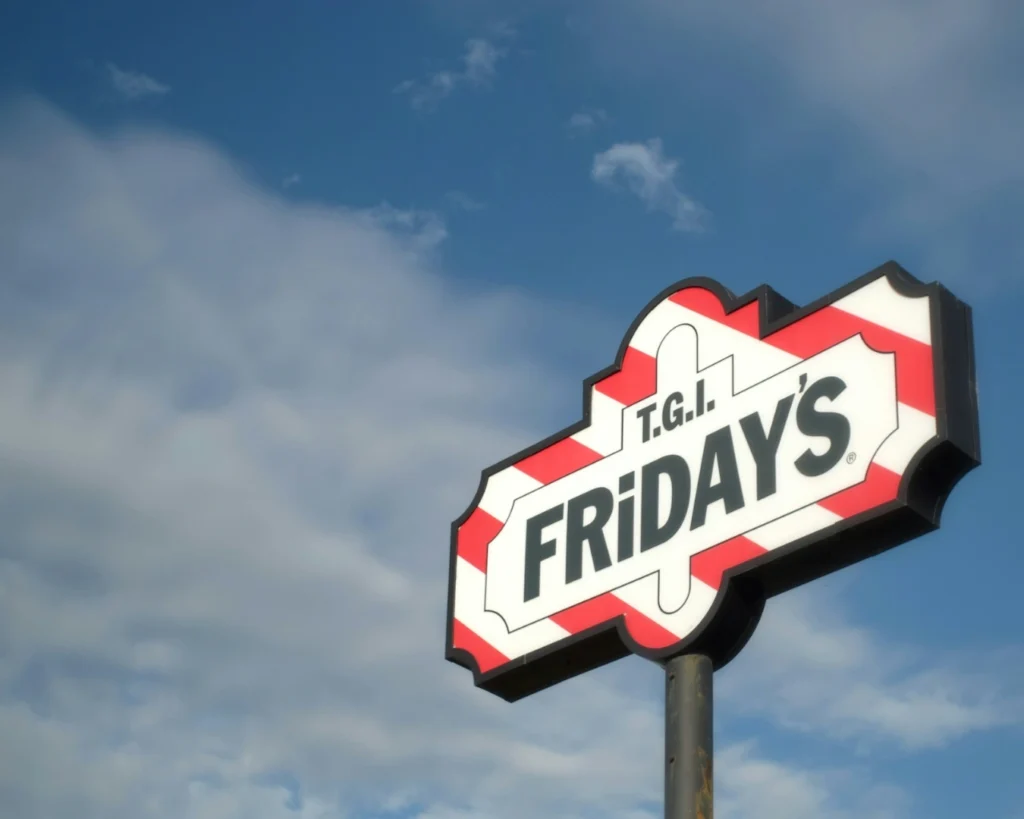 A TGI Friday's sign with bold letters is set against a partly cloudy sky. The sign has a red and white striped border and conveys a casual dining atmosphere.