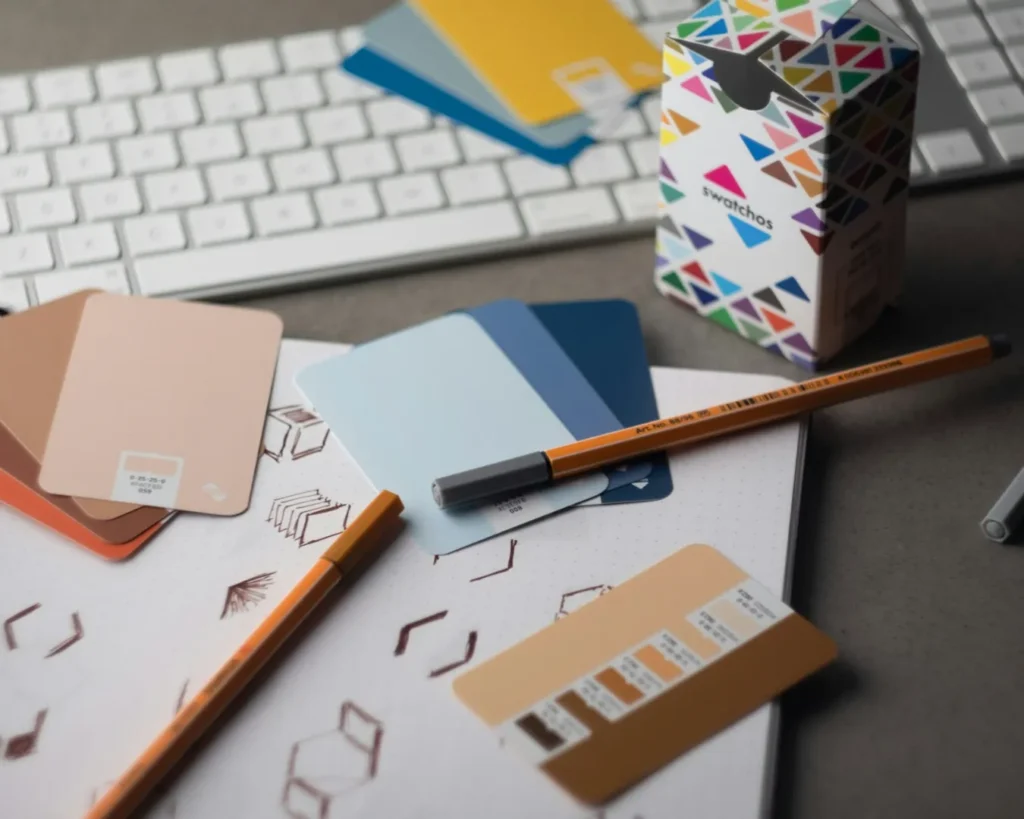 Close-up of a design workspace showing color swatches in browns, blues, and yellows on a notebook with geometric sketches, pens, and a keyboard.