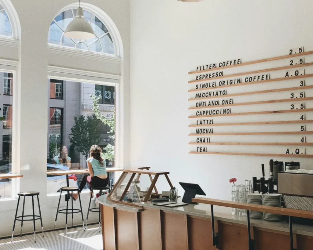 Bright coffee shop with large arched windows, a person seated at a counter, and a menu listing various coffee options on the wall. Airy and serene ambiance.