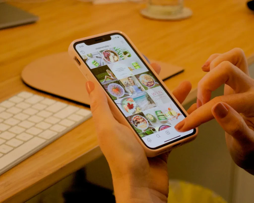 A person holds a smartphone, browsing a vibrant social media feed filled with colorful food and drink images. A keyboard is visible on a wooden desk.