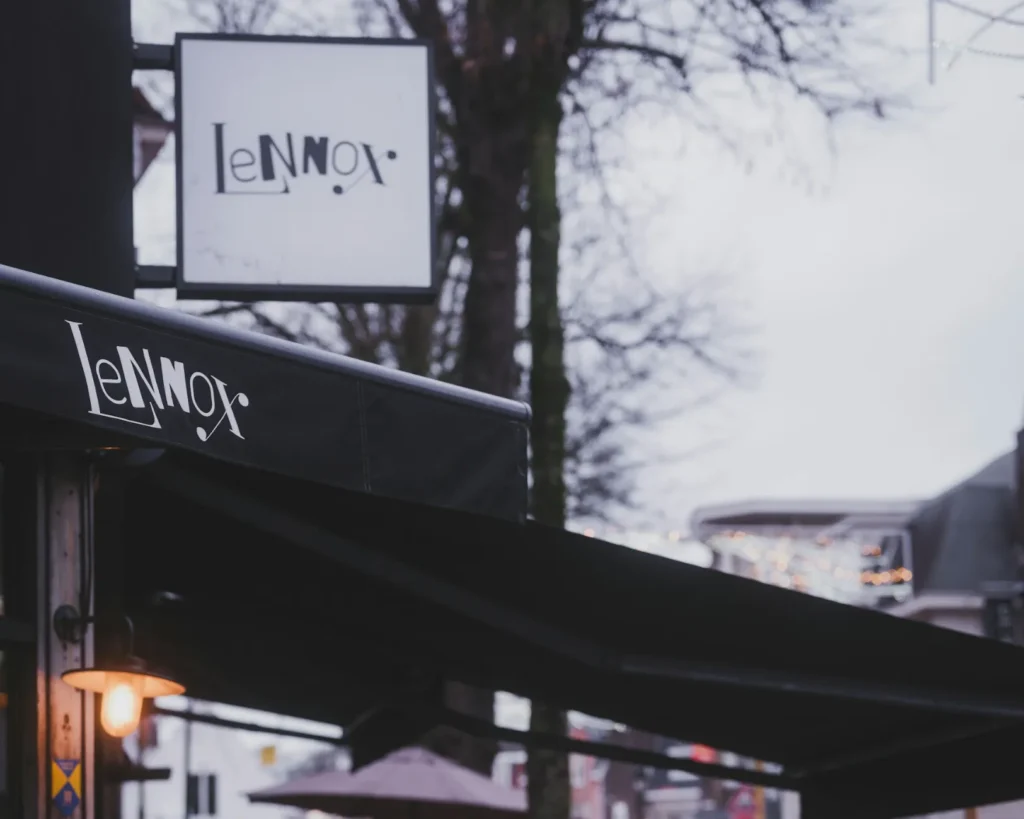 Street view showcasing a dimly lit café with "Lennox" on an awning and sign. Soft lighting under the awning creates a cozy, inviting atmosphere.