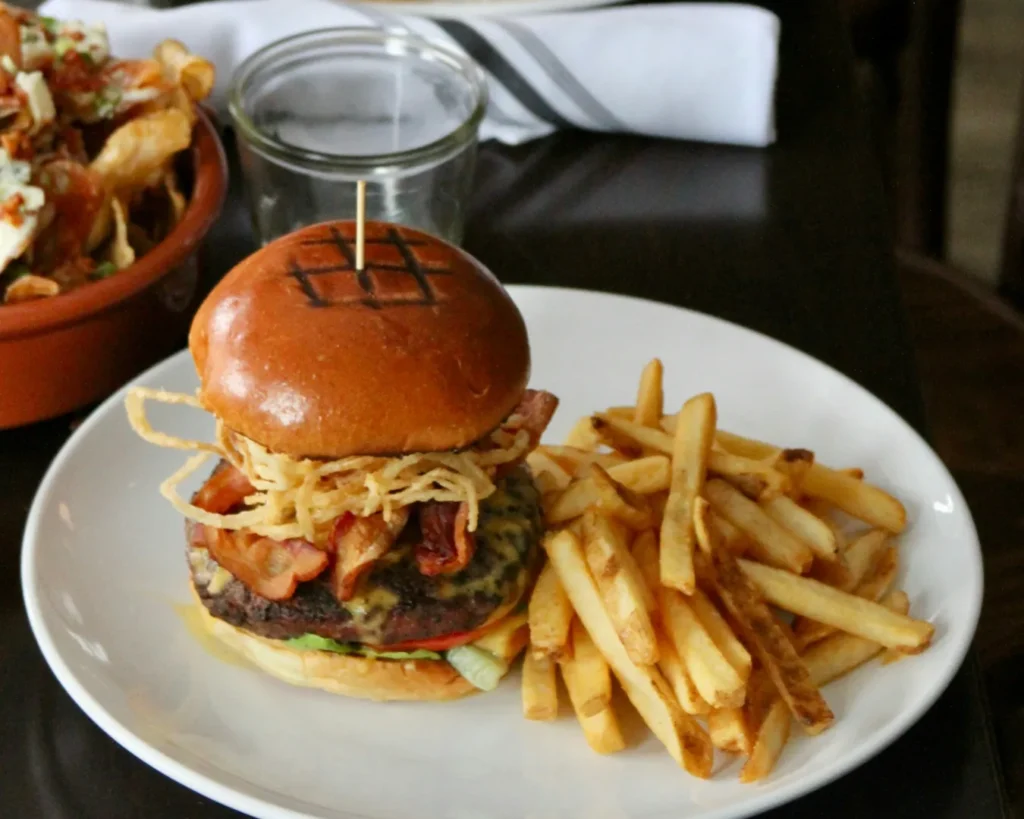A gourmet burger with crispy onions, bacon, and cheese on a toasted bun sits on a white plate, paired with golden fries; a small bowl of loaded potato skins in the background.