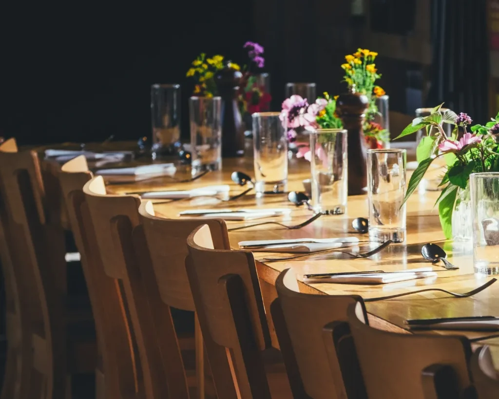A sunlit wooden table set for a meal with empty glasses, silverware, and vibrant floral centerpieces, conveying a warm, inviting atmosphere.