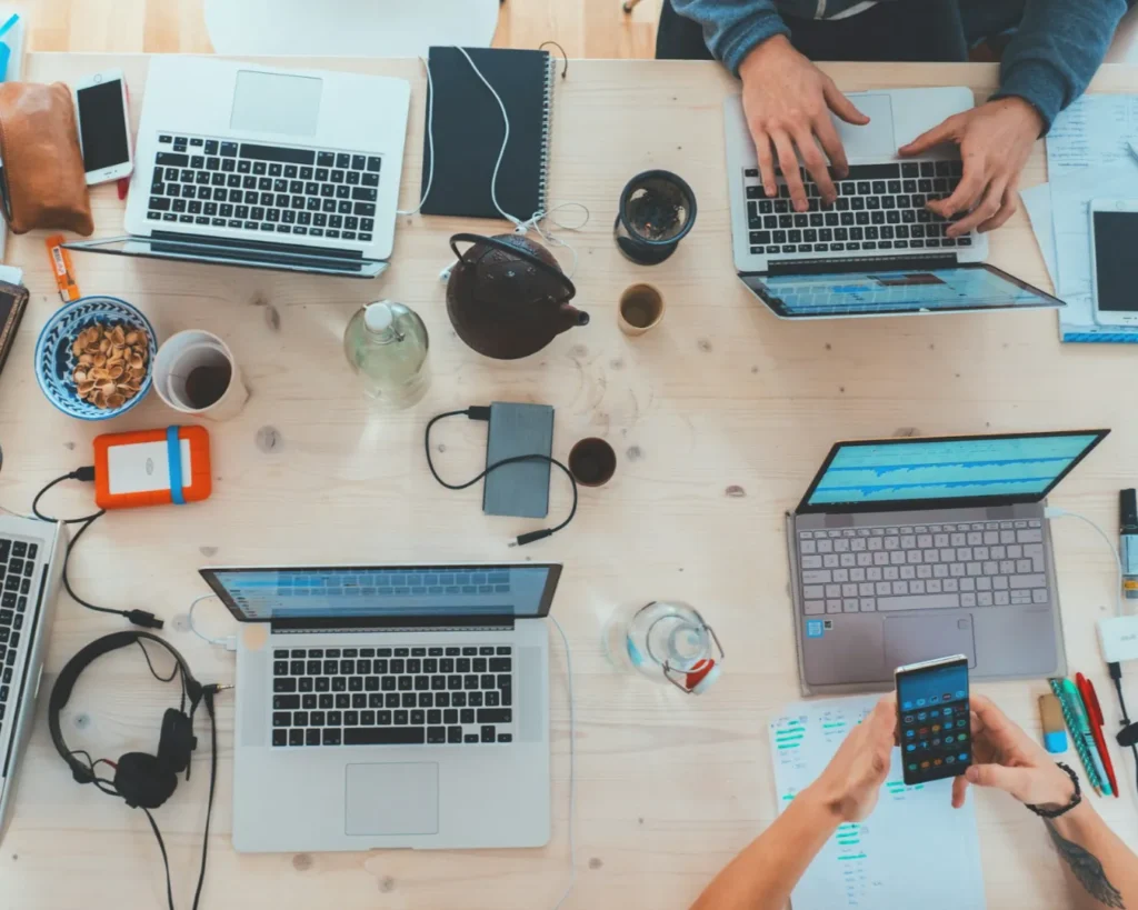 Overhead view of a cluttered desk with laptops, smartphones, notebooks, and coffee. People work collaboratively, creating a busy and productive atmosphere.