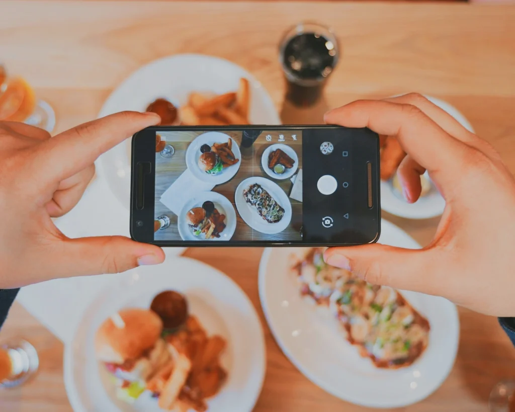 Hands hold a smartphone over a wooden table, capturing a top-view photo of assorted plates with food, including a burger and fries, conveying a casual dining vibe.