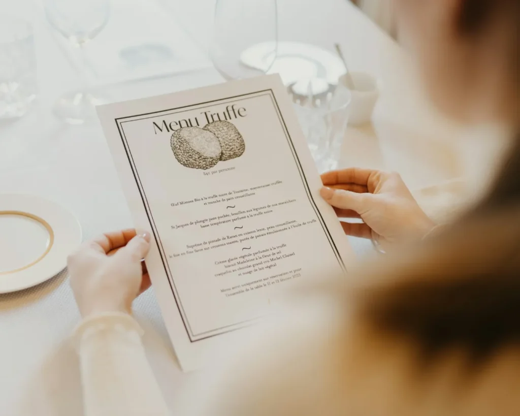 A woman holds a "Menu Truffe" at a elegant dining table, featuring glasses and a saucer. The atmosphere is refined and sophisticated.