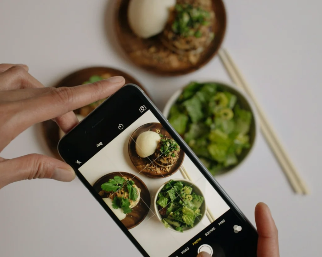 Close-up of a phone capturing food on a table, including bowls of salad, noodles, and a bun. Chopsticks are nearby, evoking a casual dining scene.