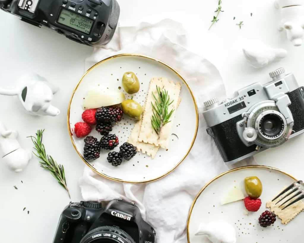 Aerial view of a stylish food setup with two plates of berries, olives, and crackers, surrounded by vintage cameras and ceramic animal figurines on a white background.
