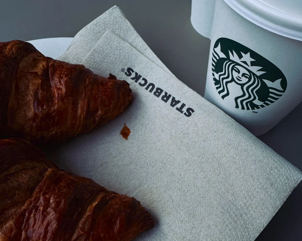 A warm, flaky croissant rests on a Starbucks napkin beside a white Starbucks coffee cup with the green logo, creating a cozy café vibe.