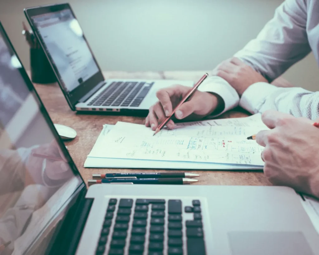 Two individuals collaborate at a wooden desk, reviewing notes on a paper with laptops open. The scene conveys teamwork and focus.