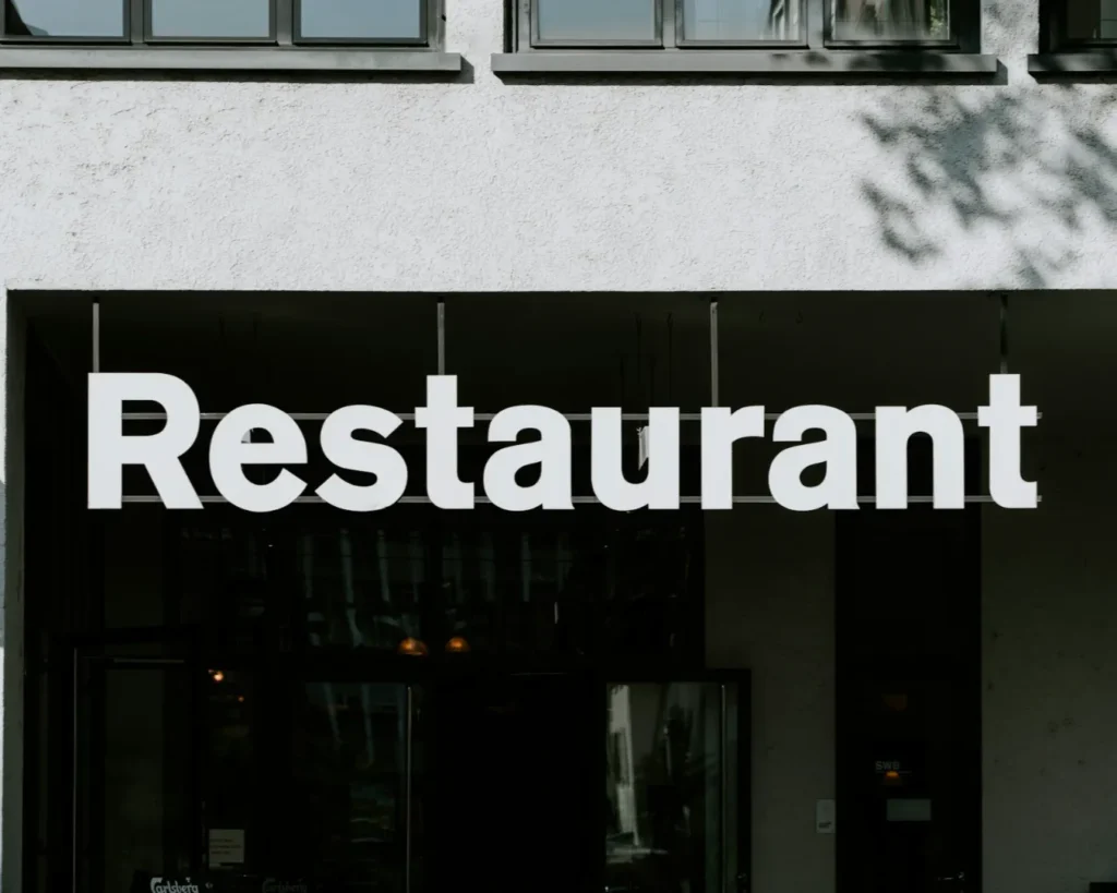White "Restaurant" sign on a building facade with gray walls and large windows. The tone is modern and minimalistic, evoking a welcoming atmosphere.