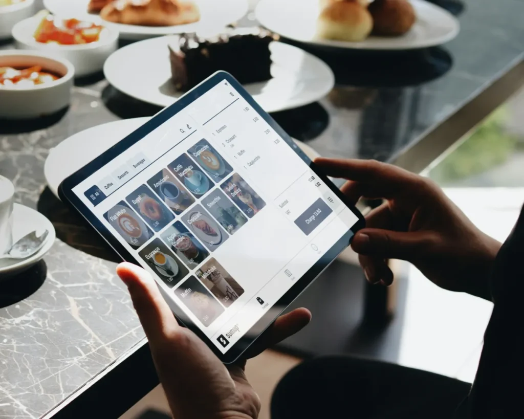 A person holds a tablet displaying a digital menu, with food and drink options, at a restaurant table set with plates of pastries and drinks. The atmosphere is modern and casual.
