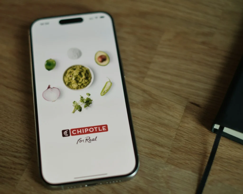 Smartphone on wooden table displays Chipotle app screen with ingredients like guacamole, avocado, onion, and chili. Chipotle logo and "for Real" text visible.