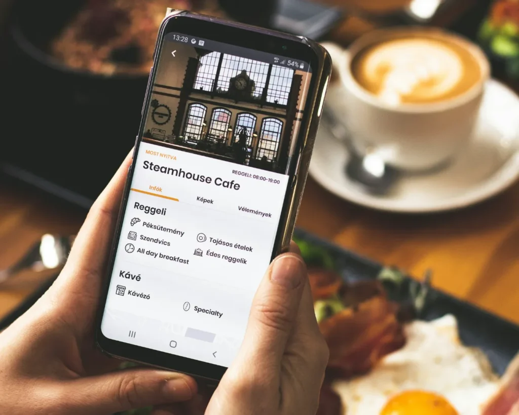 A person holds a smartphone displaying the Steamhouse Cafe menu app, showing breakfast and coffee options. A cappuccino and breakfast plate are in the background.