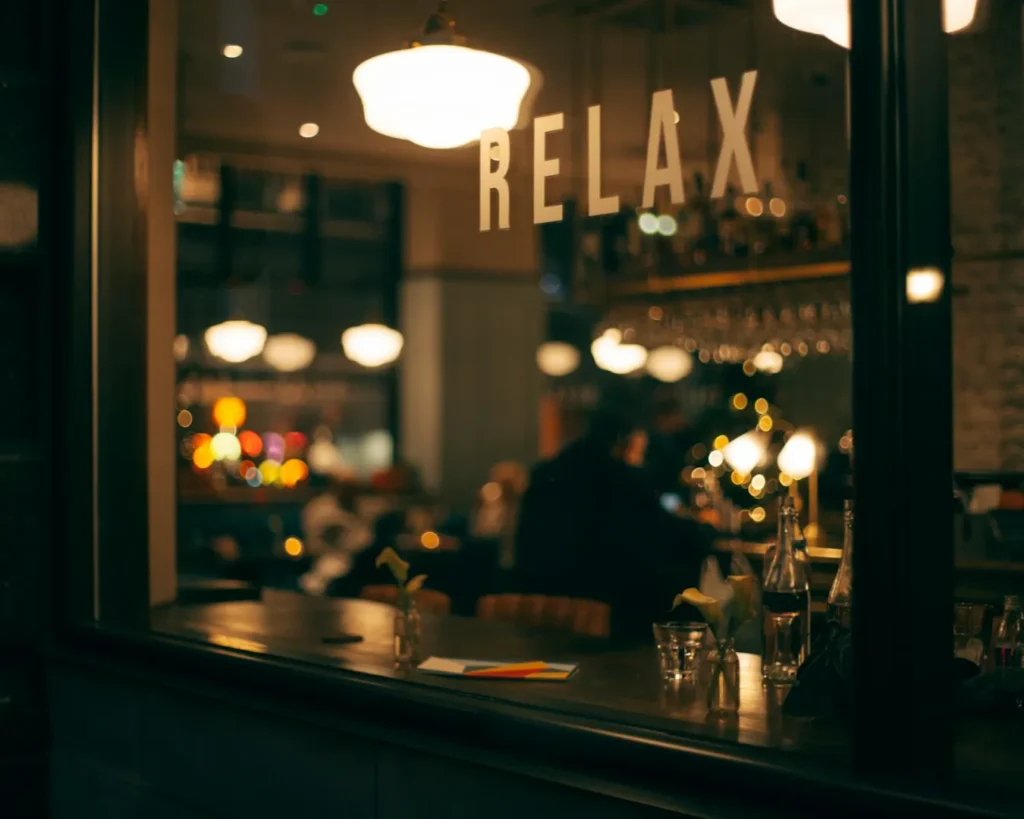 Cozy cafe interior viewed through a window with "RELAX" on the glass. Warm lighting, blurred figures inside, conveys a calm, inviting ambiance.
