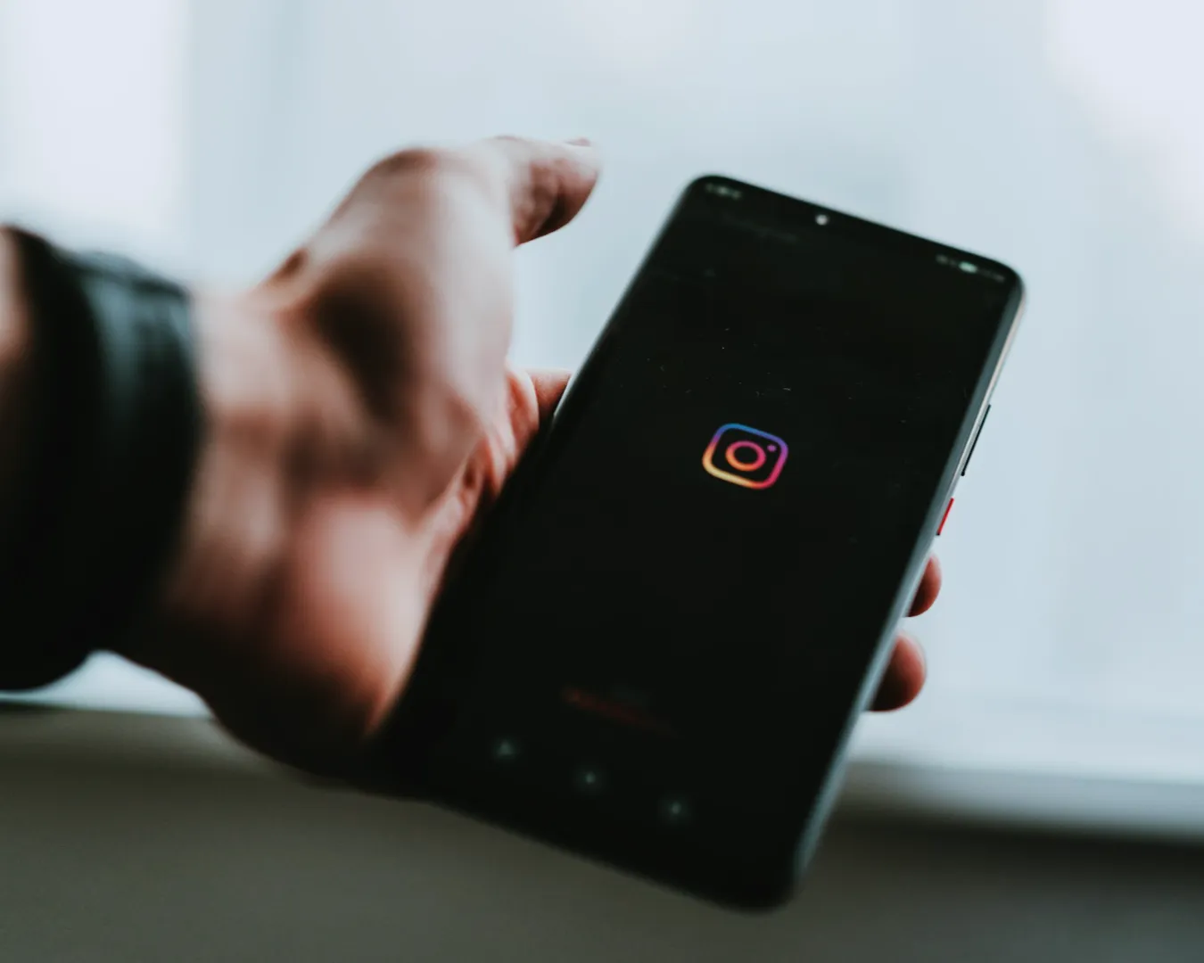 A hand holds a smartphone displaying the Instagram logo on a black screen. The background is softly blurred, creating a focused and modern feel.