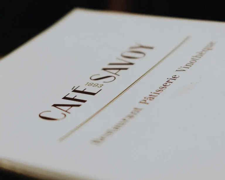 Close-up of a Café Savoy menu with elegant serif font highlighting "Café Savoy 1893." The text suggests a vintage and classy atmosphere.
