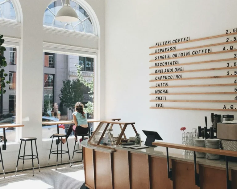 Bright café with high ceilings, white walls, and large arched windows. A menu board lists drinks. A person sits by the window. Modern, airy vibe.