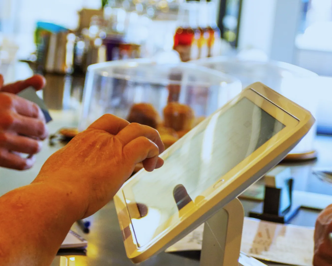 Hands interact with a touchscreen register in a cafe, with pastries under glass domes and blurred bottles in the background, creating a busy, modern vibe.