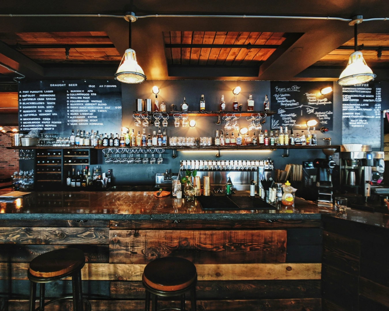 Cozy bar interior featuring a wooden counter with stools, hanging lights, and shelves stocked with bottles. Chalkboard menus display drink options. Warm, inviting ambiance.
