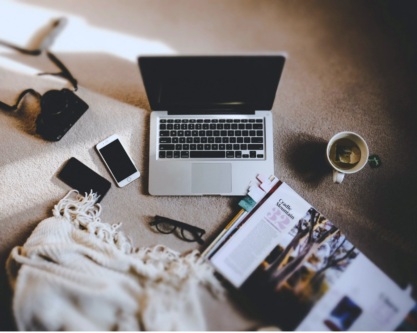 Cozy workspace on a carpet featuring an open laptop, smartphone, camera, magazine, reading glasses, a fringed blanket, and a cup of tea.