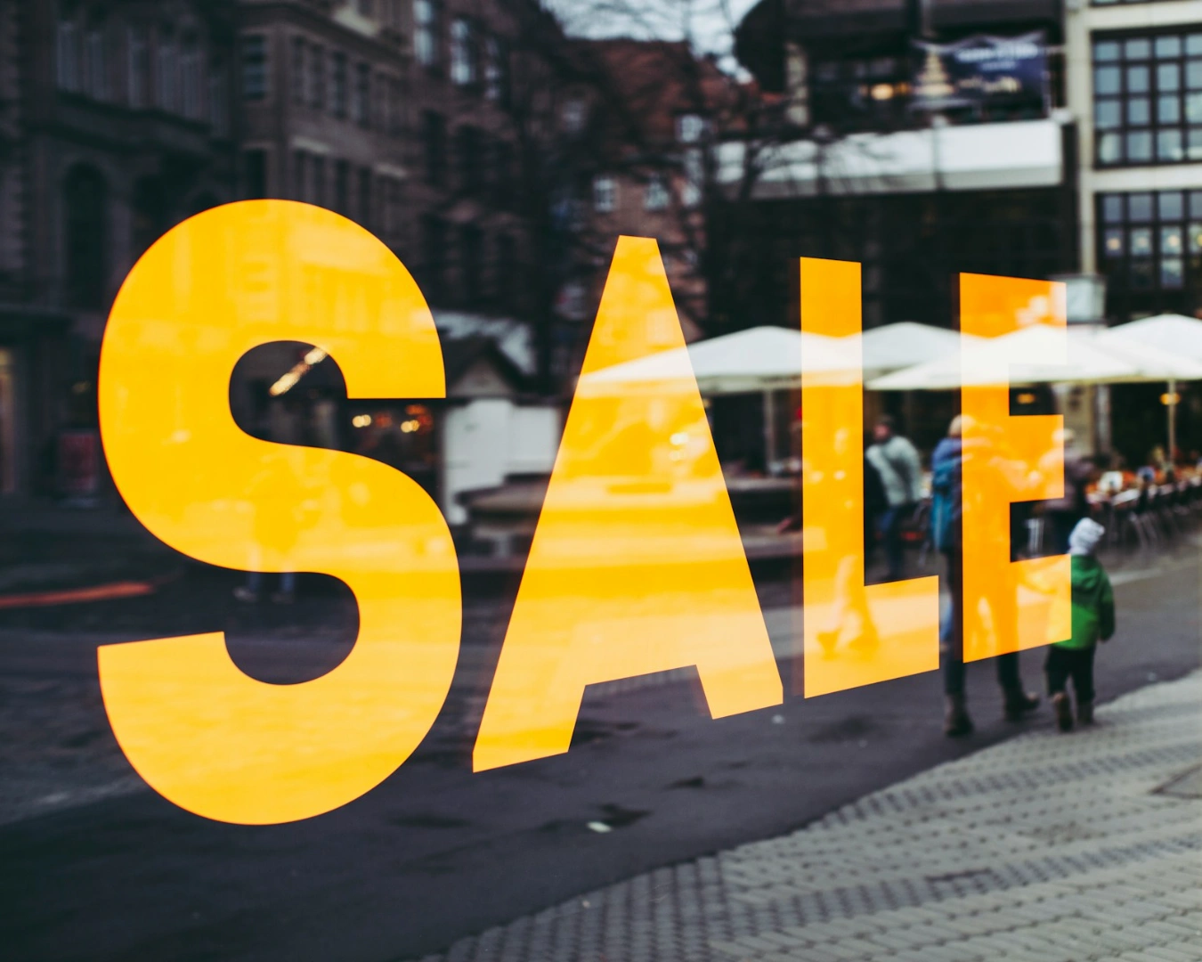 Yellow "SALE" text on a shop window, reflecting a busy street scene with people and outdoor seating. The atmosphere is lively and bustling.