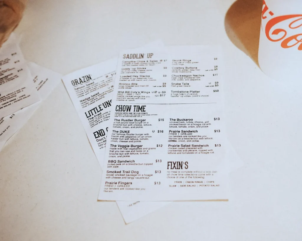 A restaurant menu on a beige table, displaying various burger and sandwich options with prices. A paper cup with partial "Coca-Cola" logo is visible.