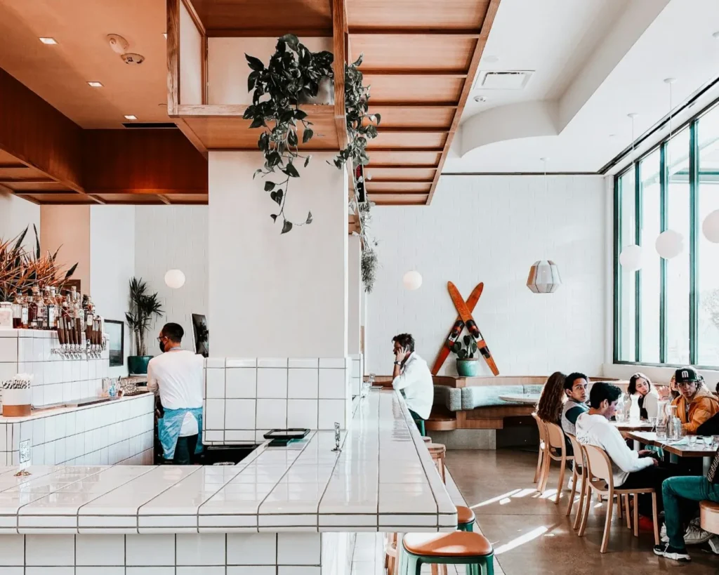 Bright café interior with modern decor. A barista works behind a tiled counter, while a group of people sit at a table, chatting and enjoying drinks.