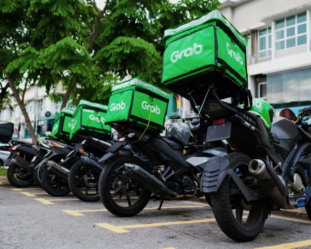 A row of parked motorcycles with bright green Grab delivery bags, under leafy trees, in an urban area. The scene conveys readiness and efficiency.