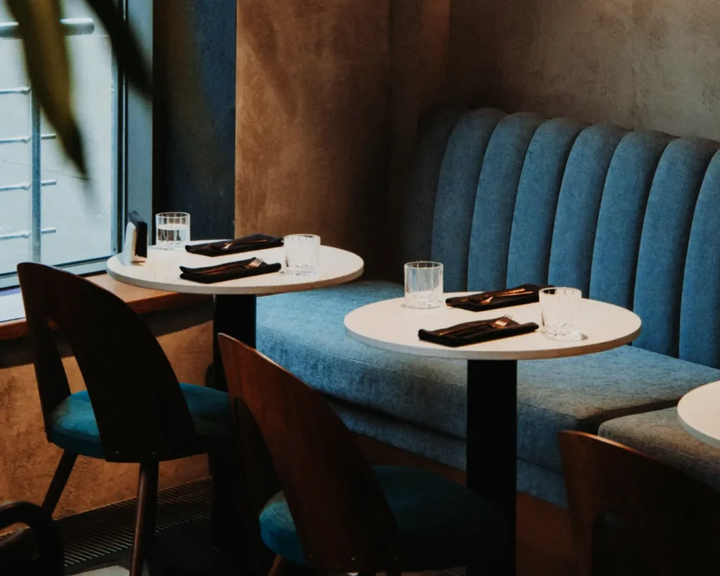 Cozy cafe interior with two round tables set for dining, featuring blue cushioned booth seating, dark chairs, napkins, and glasses near a window.