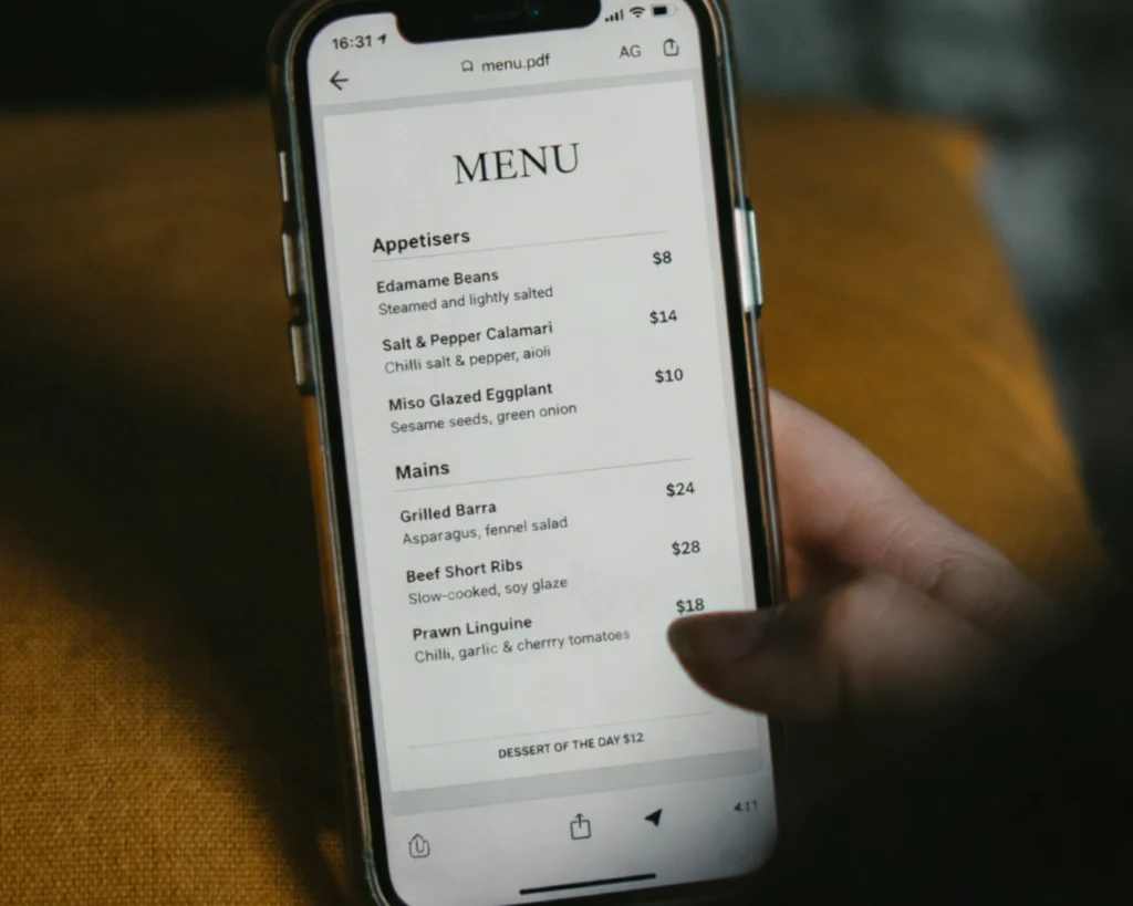 A hand holds a smartphone displaying a restaurant menu with items like edamame beans, calamari, and beef short ribs. The setting is cozy and inviting.