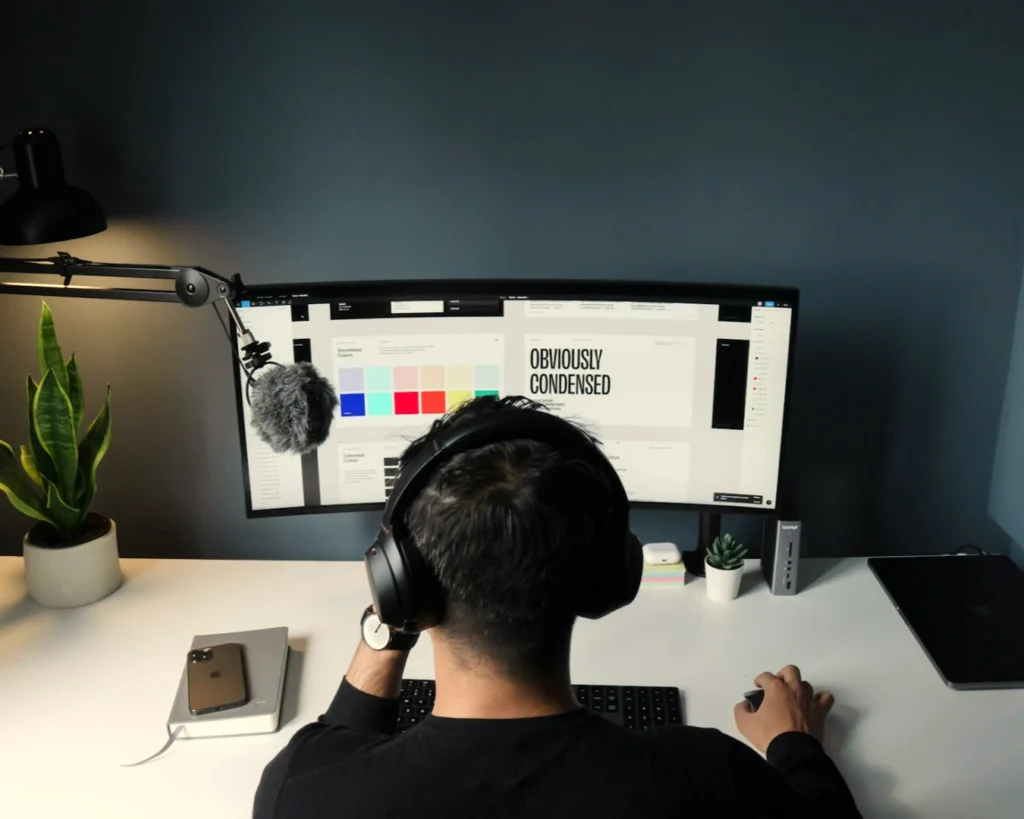 A person wearing headphones works at a dual-monitor setup displaying design software. A microphone, smartphone, and potted plant are on the white desk.