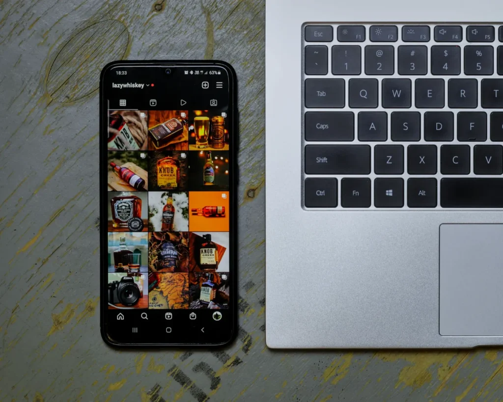 A smartphone displaying a whiskey-themed Instagram profile is placed beside a silver laptop on a wooden surface, creating a tech and lifestyle vibe.