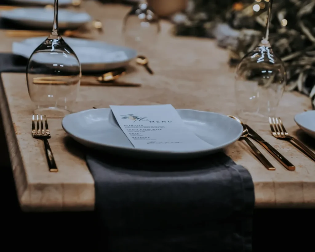 Elegant table setting with a white plate, golden cutlery, and a wine glass. A menu rests on the plate atop a gray napkin, evoking sophistication and warmth.
