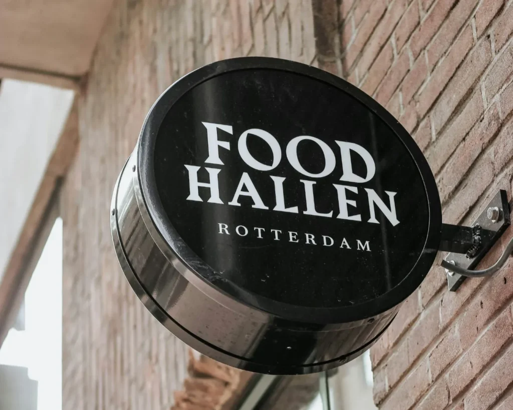 Round black sign with white text reads "FOOD HALLEN ROTTERDAM," mounted on a brick wall. The setting appears urban and modern, conveying a stylish vibe.