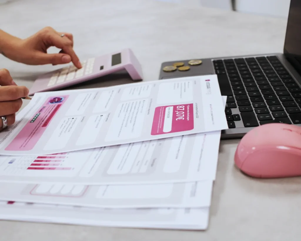 Hands using a pink calculator with printed documents, a laptop, and a pink mouse on a desk, conveying a focus on budgeting and finance.