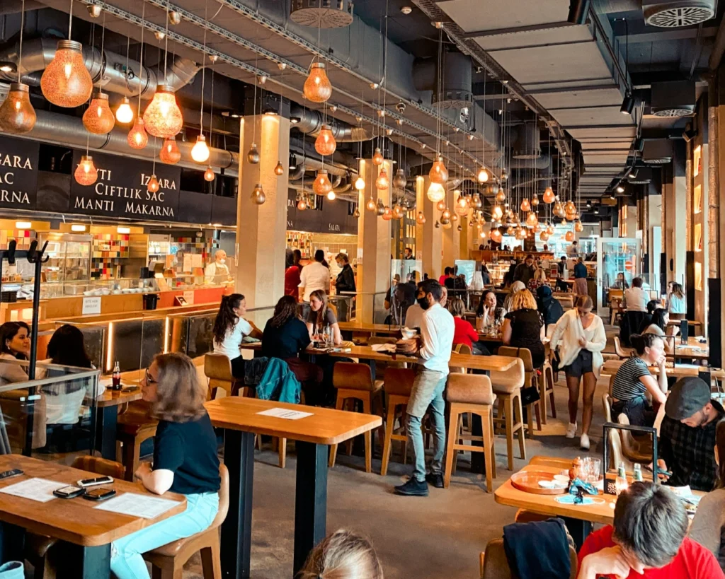 Lively restaurant with a modern industrial design features hanging lights, wooden tables, and patrons enjoying meals. Warm, inviting atmosphere.