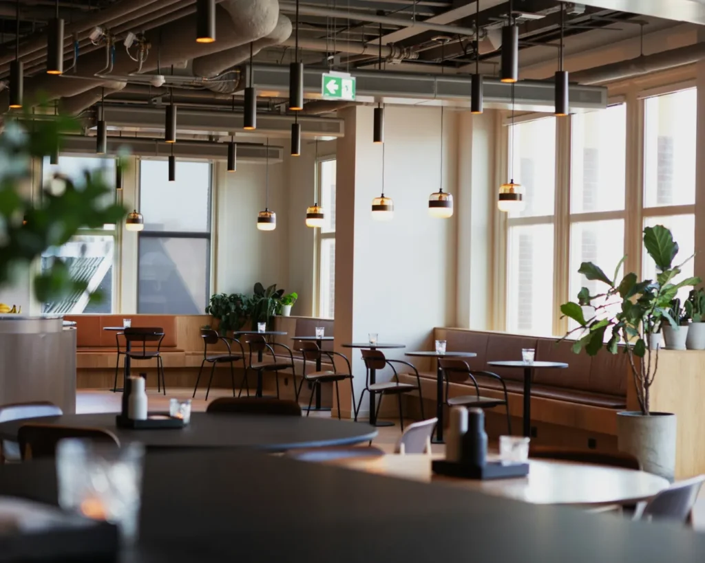 Modern cafe interior with industrial design, featuring black pendant lights, wooden seating, and large windows. Lush green plants add a cozy touch.