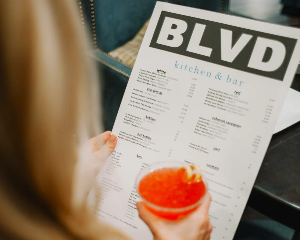 A person views a menu from BLVD Kitchen & Bar while holding a vibrant red cocktail. The tone is relaxed and inviting, suggesting a dining experience.
